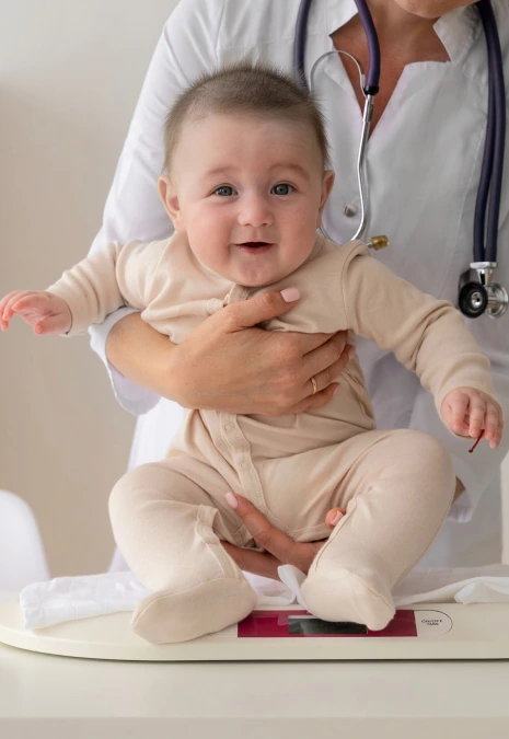 Muayene masasında beyaz önlüklü sağlık uzmanının kucağında oturan gülümseyen bebek